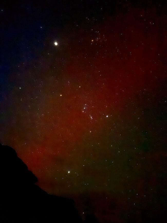 Red and green aurora behind the constellation Orion, with a rock outcrop silhouetted at the edge