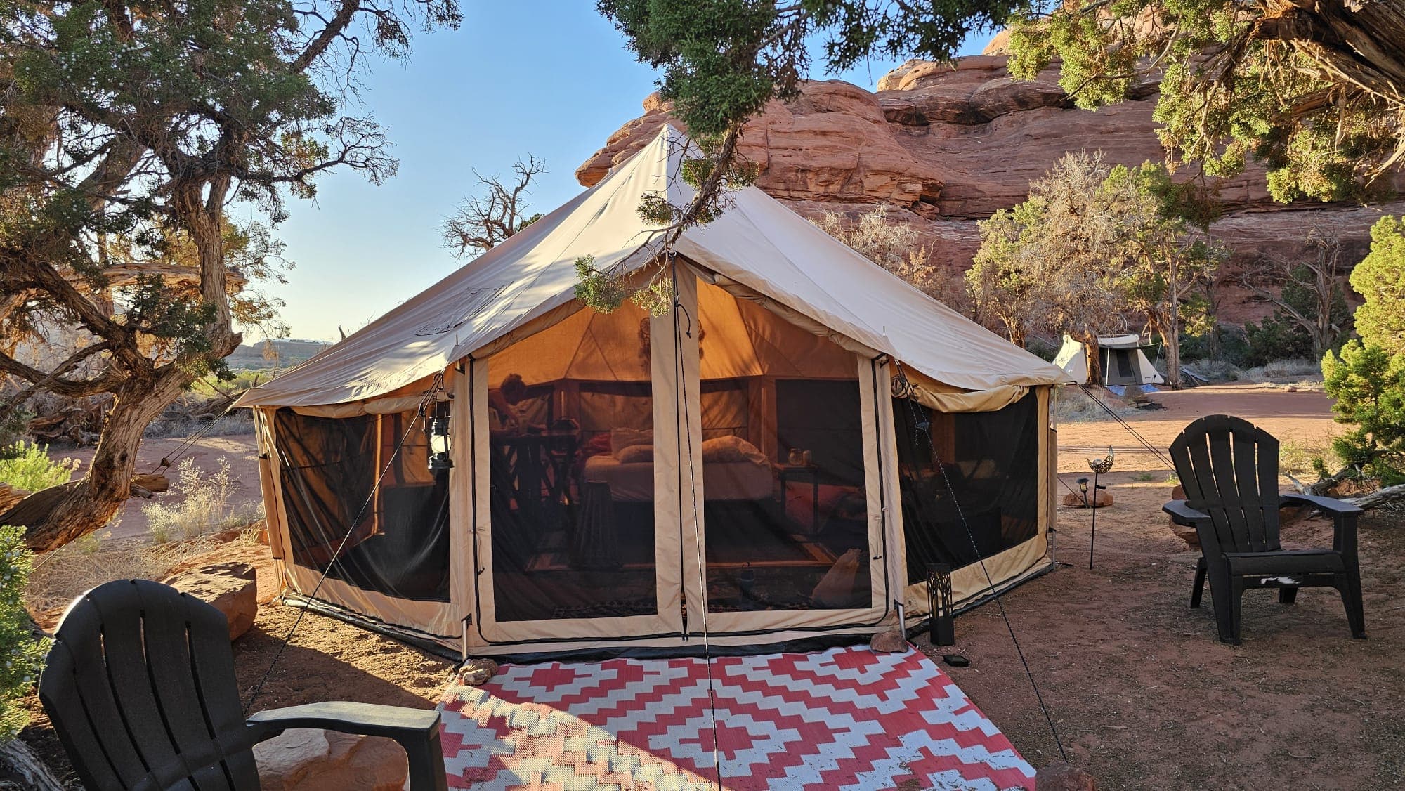 The Mo'Rockin Bell Tent lit from within at golden hour, under juniper with a red rock wall behind