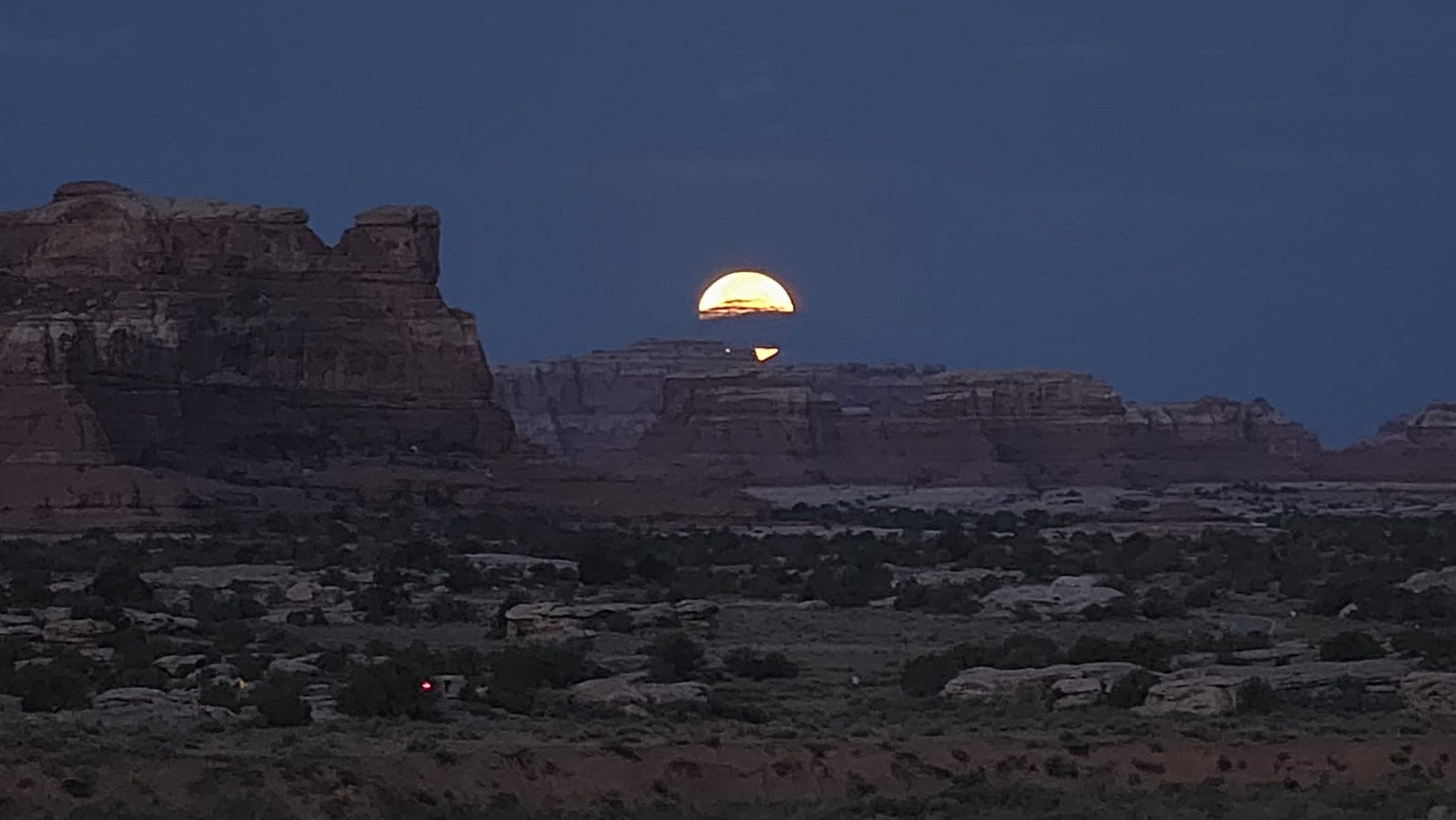 A deep orange full moon setting behind the layered sandstone of the Needles at dusk