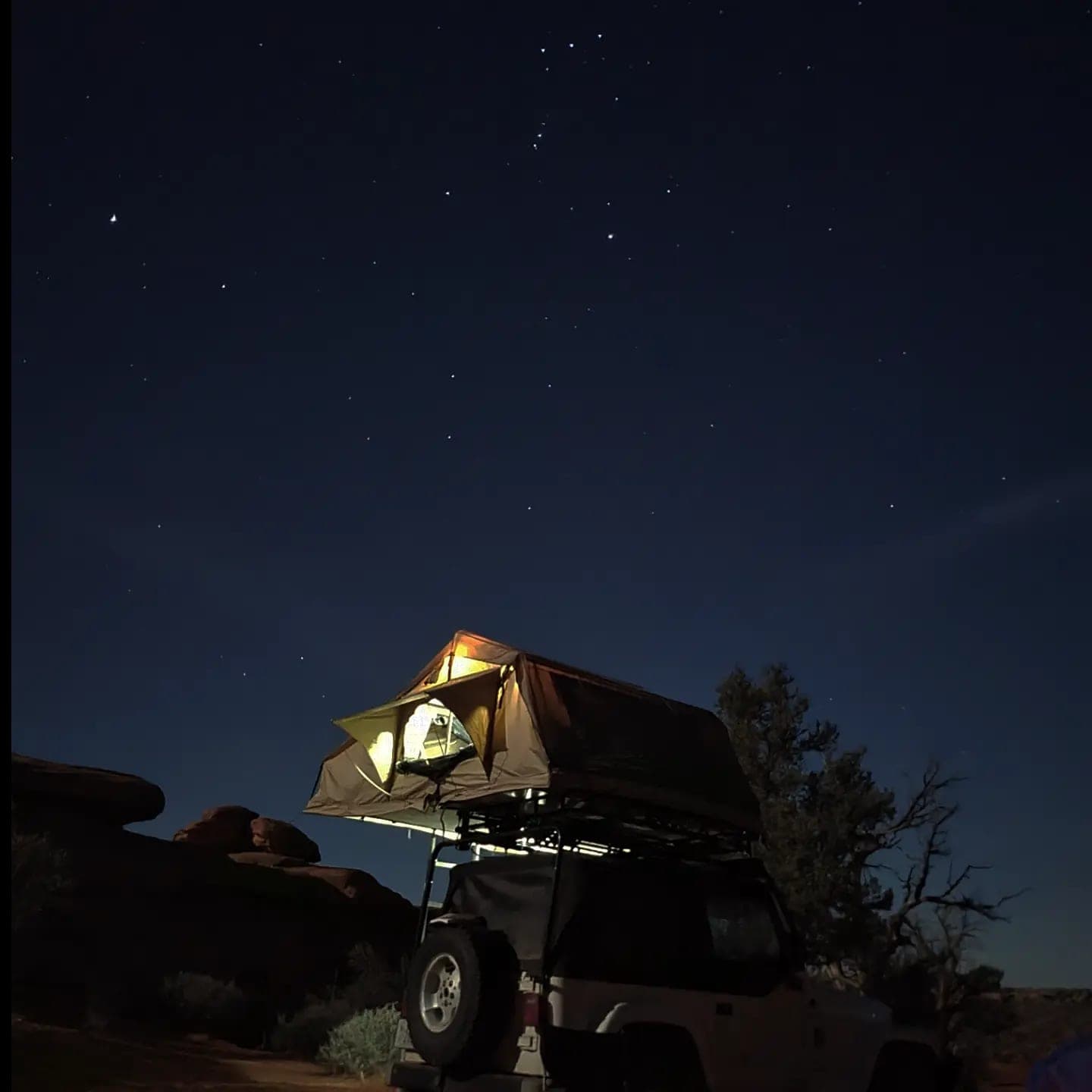 A rooftop tent on a vehicle, lit warm from inside, under a star-filled night sky beside a juniper